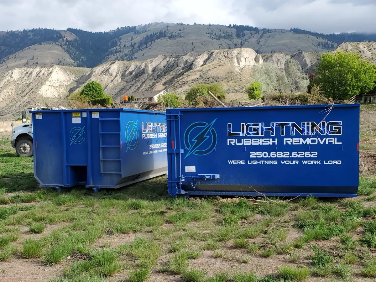 Two blue Lightning Rubbish Removal roll-off bins on grass with the Kamloops hills behind
